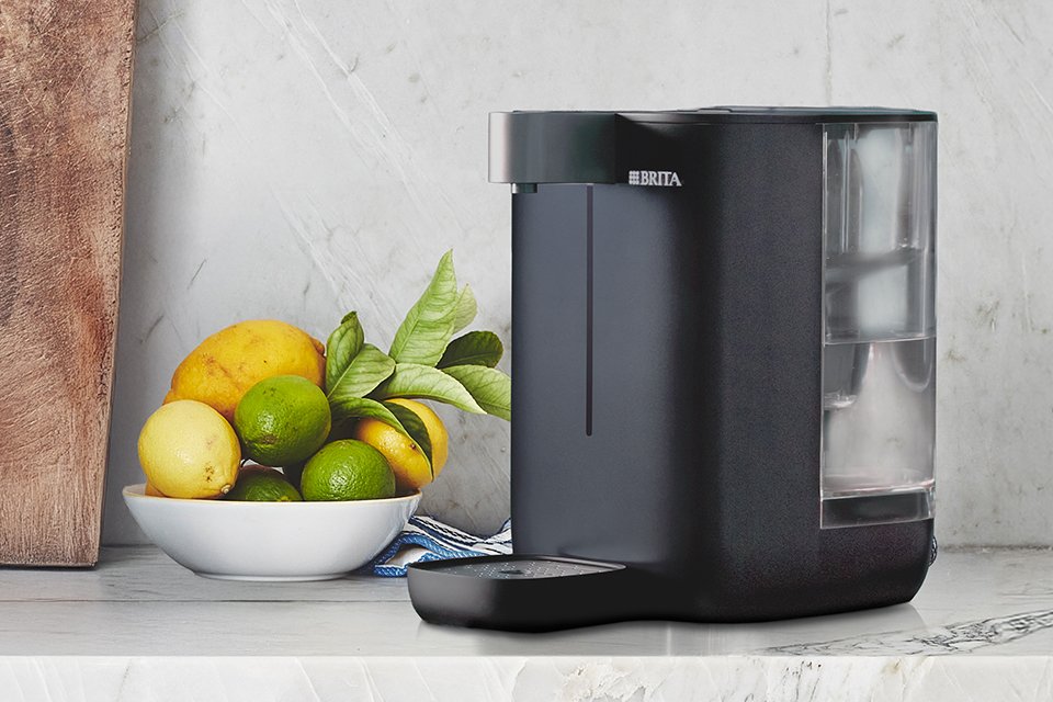 A BRITA YOURCE mini ambient and hot water dispenser on a kitchen worktop next to a bowl of fruit.