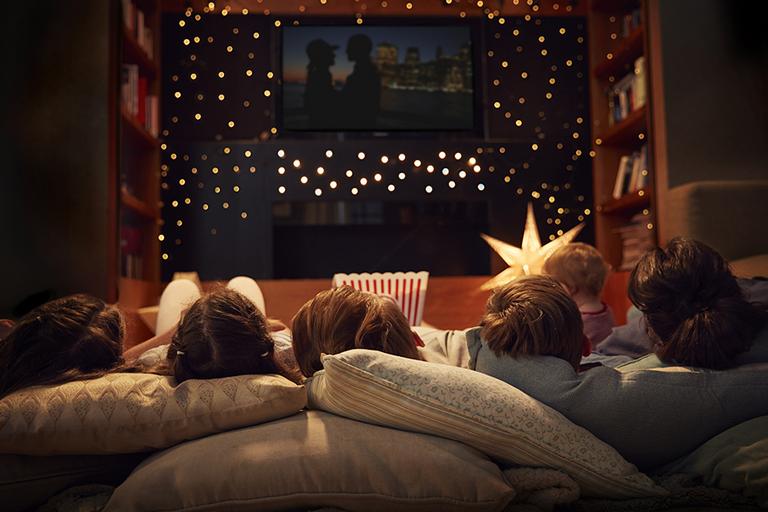 Family led on cushions on the floor, watching a film on a TV screen surrounded by fairy lights.