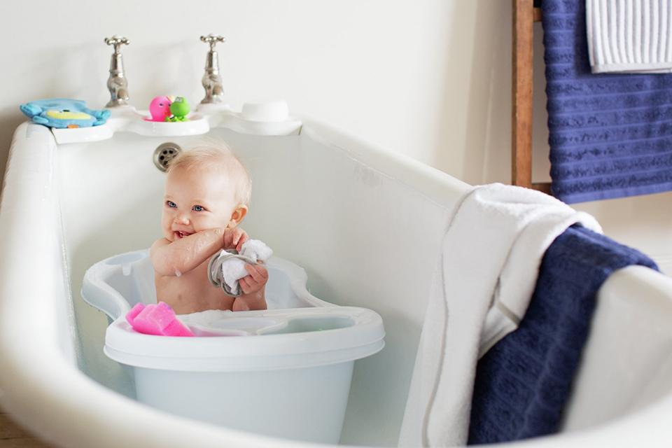 A smiling baby is shown sitting in a baby bath, with fluffy towels and bath toys nearby.