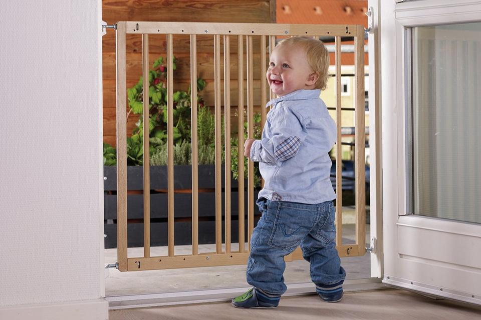 A toddler is standing in front of a baby gate, installed in an open doorway through which blue sky and neighbouring houses can be seen.