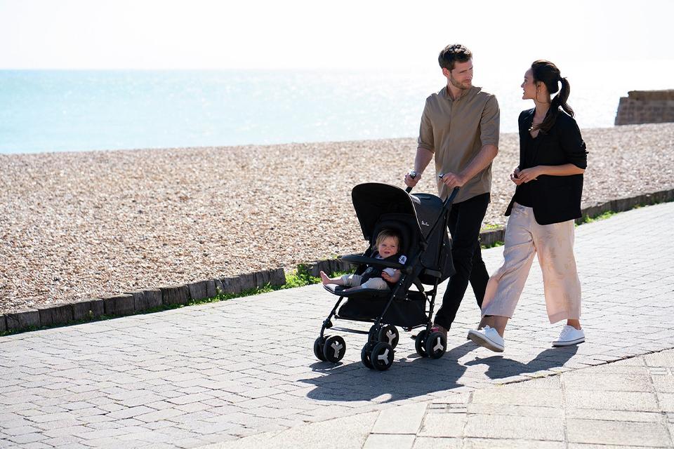 A couple stroll along a beachfront path, pushing a baby in a pram.