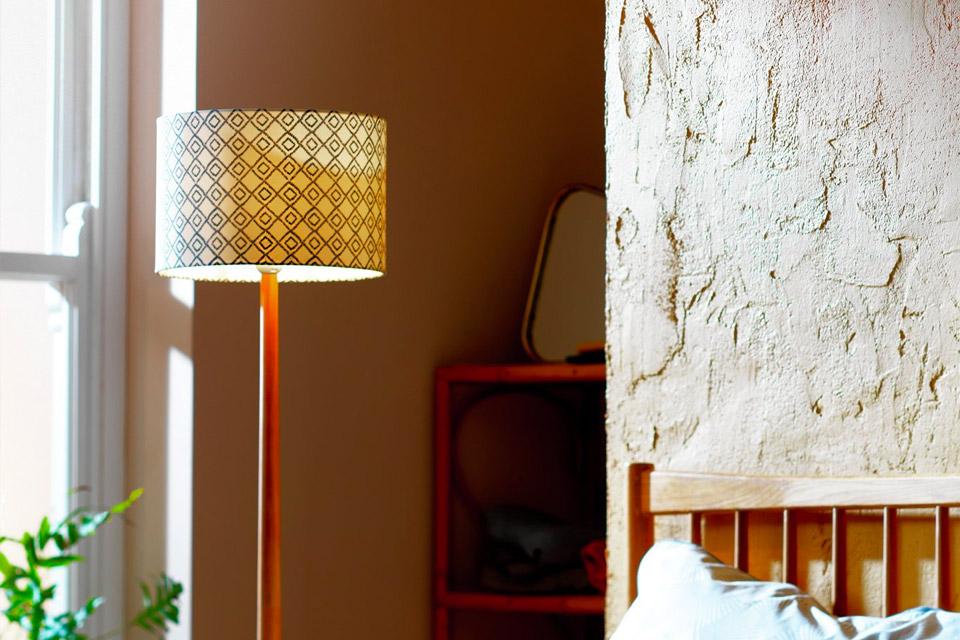 A boho style print shade on a floor lamp with a wooden base placed next to a bed. 