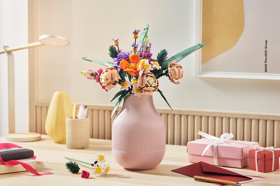 Image shows a bouquet of Lego flowers arranged in a pink vase, next to gift boxes and envelopes on a wooden table.