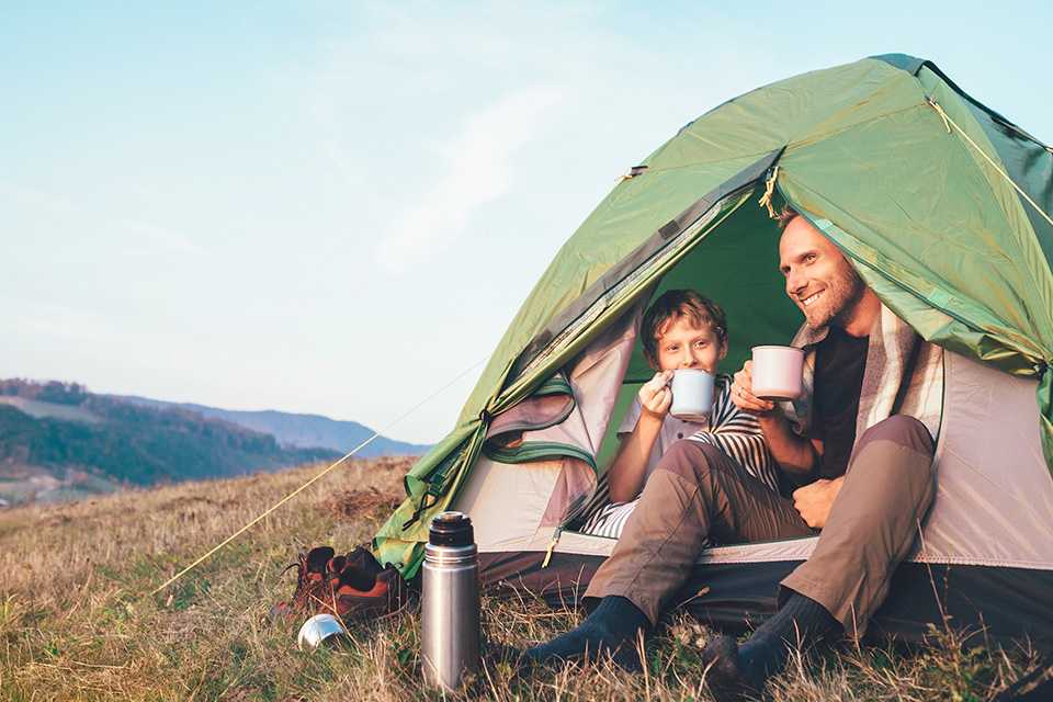 A father and son sip tea in the entrance to a green tent, set up on the slopes of a tall hill.