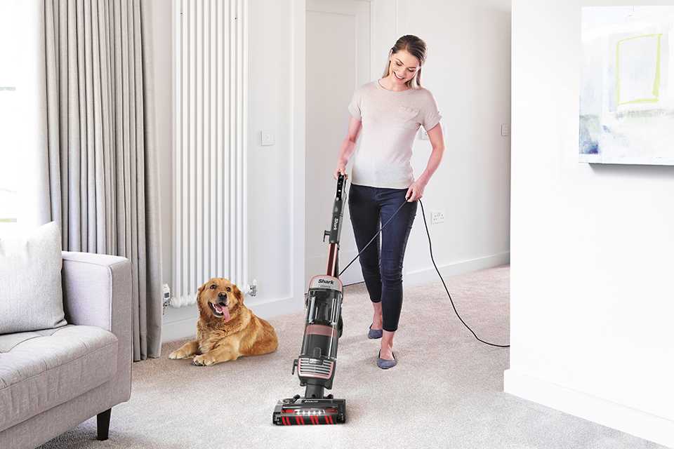 A dog sitting next to a woman cleaning the floor using a corded Shark vacuum cleaner.