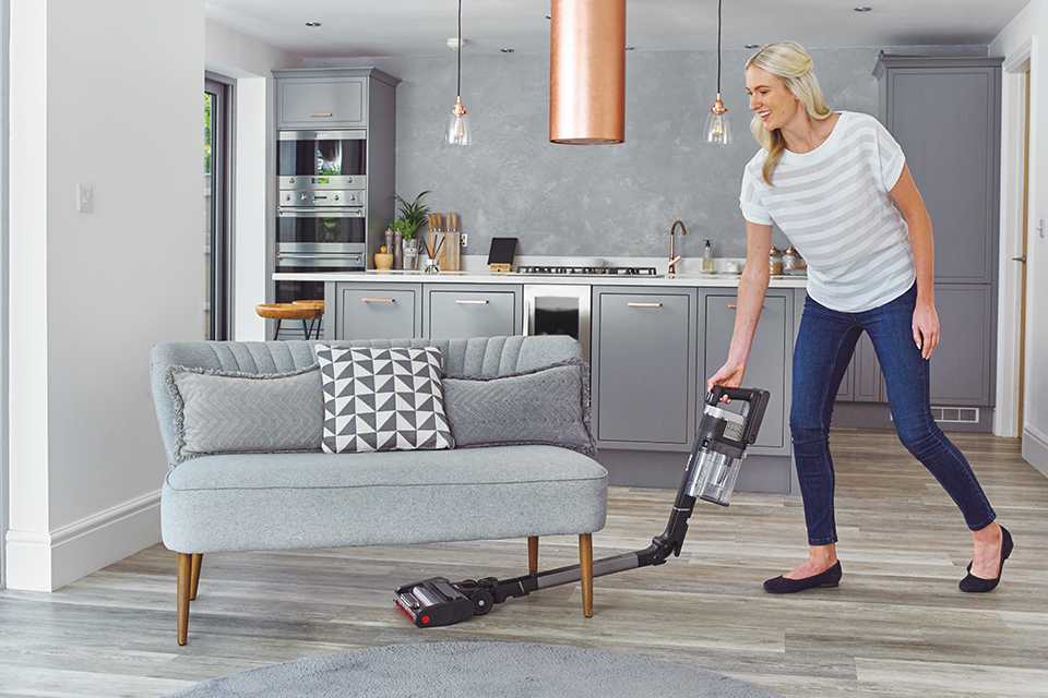A woman using a cordless Shark vacuum cleaner to clean the floor under a sofa.