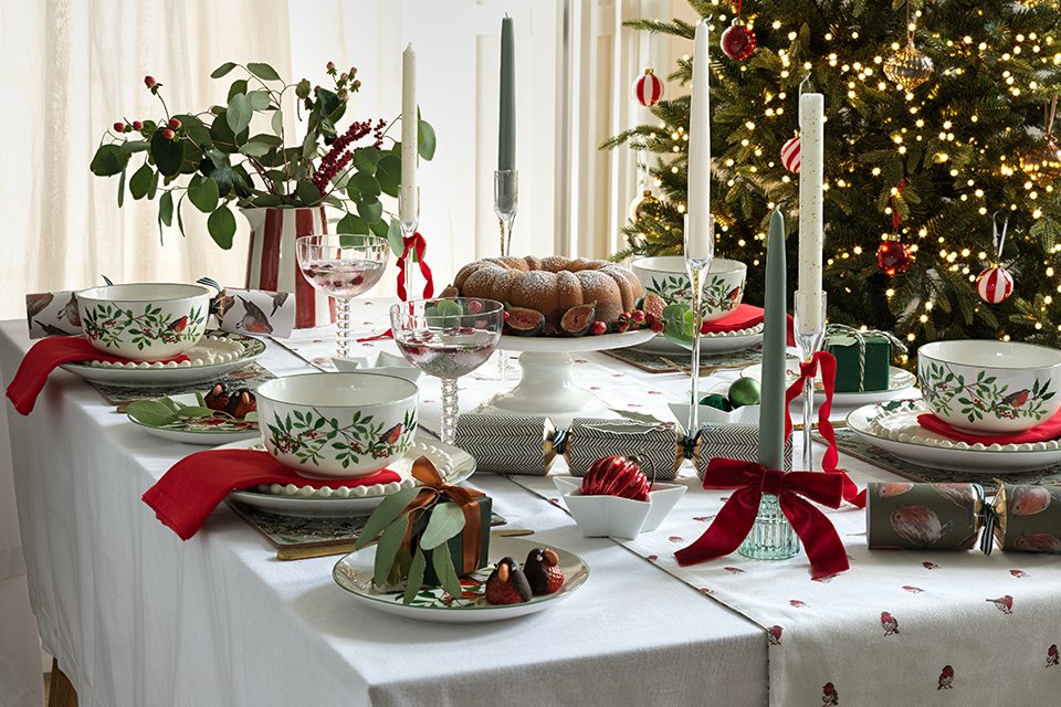 White table cloth on dining table with red robin print tabelware and an array of different height candles with christmas tree in background.