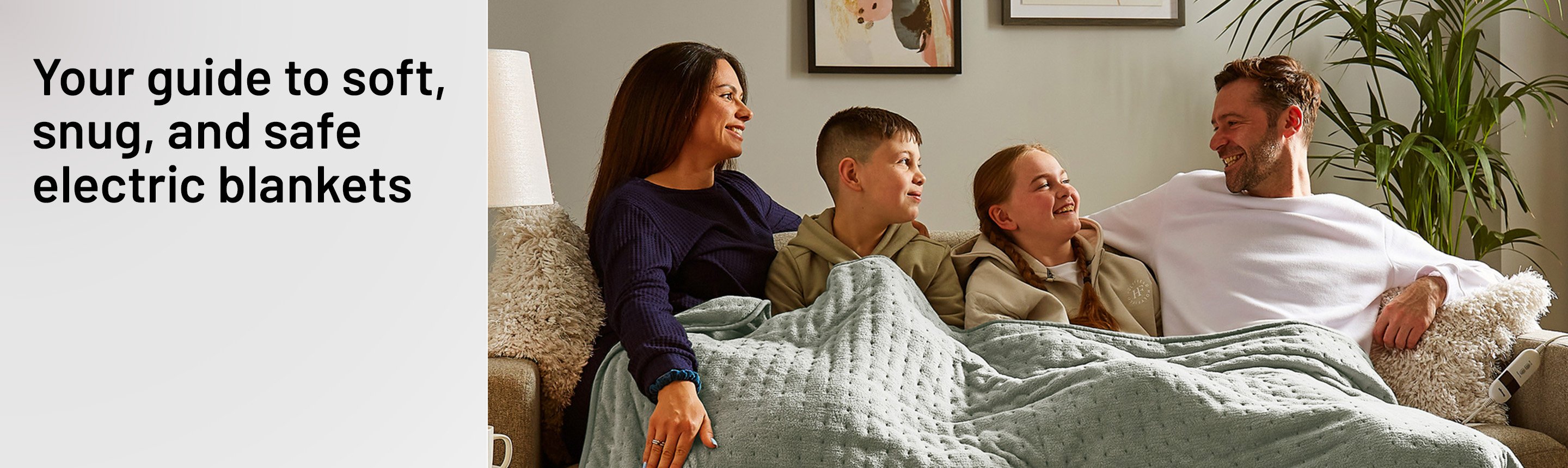 A family of 4 sitting on a sofa with Silentnight Luxury Heated Throw in sage green colour draped over them.