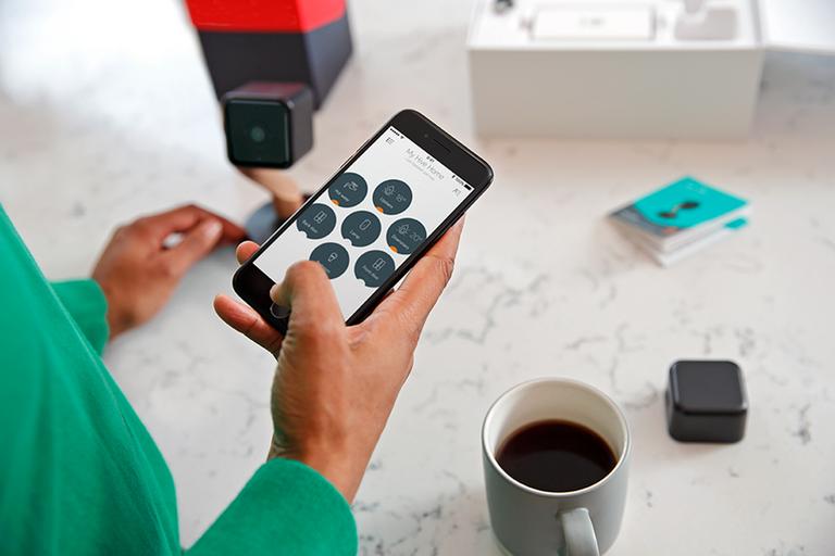 Image shows a person holding a phone, controlling different devices with the Hive app. Also in frame are a security camera and cup of coffee.