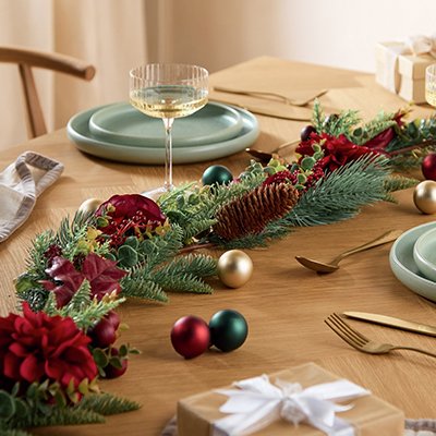 A wooden dining table set for a Christmas meal with wine glasses, cutlery, baubles, garland, dinner set and other tableware items.