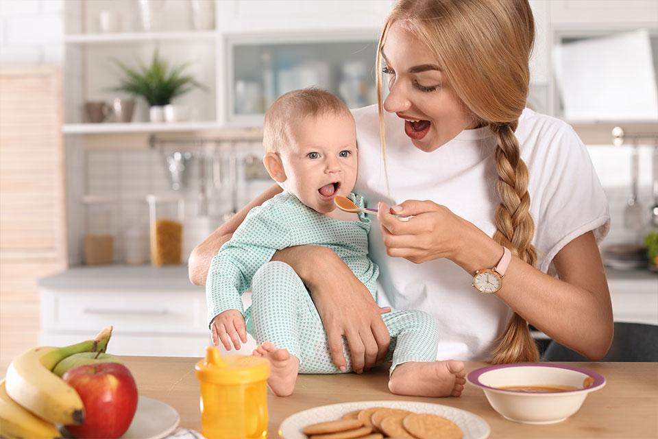 A mother feeding her baby sitting on a dining table using a spoon.