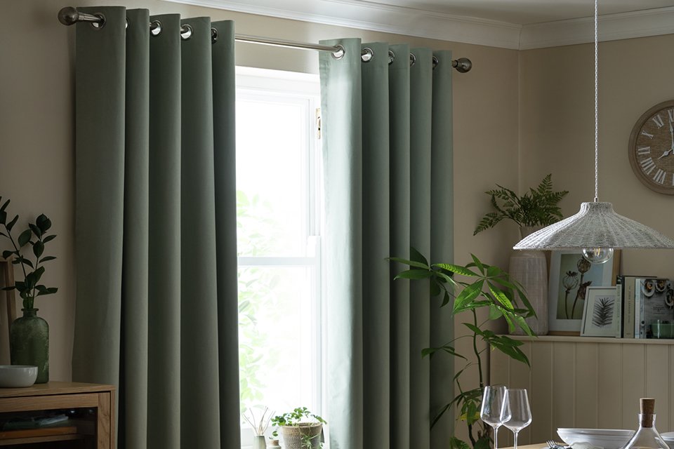 Stylishly hung sage green blackout eyelet curtains from Habitat, displayed against a white wall with natural light filtering through. The curtains feature a smooth, plain texture and silver metal eyelets, offering a modern and minimalist look.