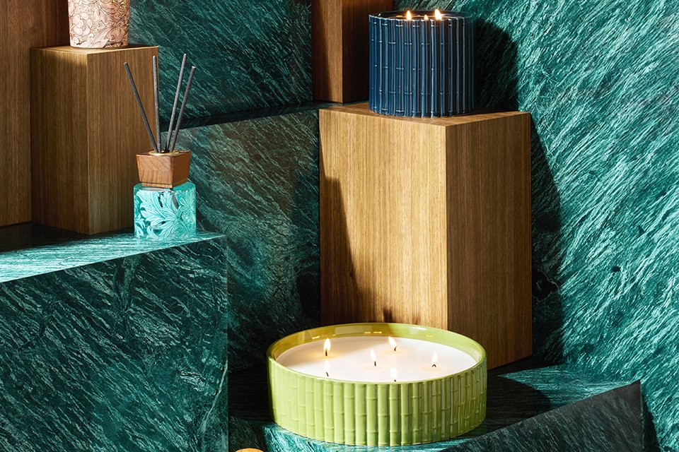 A stylish home fragrance display featuring two decorative candles and a reed diffuser on wooden pedestals, set against a green marble backdrop. The candles include a pink floral-patterned container and a dark blue ribbed container with three lit wicks, while the diffuser has black reeds in a wooden holder atop an aqua glass base with floral accents.