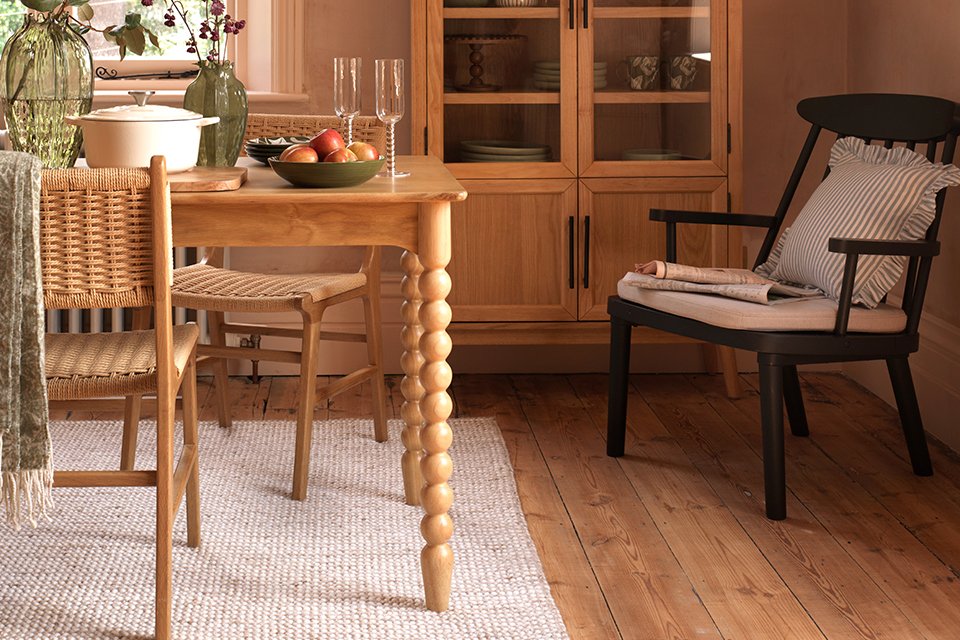Dining room with wooden furniture, featuring a round table set with a bowl of apples, glassware, and a white pot. A wicker chair and black armchair with striped cushion are placed around the table. A large arched wooden cabinet filled with dishes and glassware stands against the wall. Greenery in glass vases decorates the windowsill.