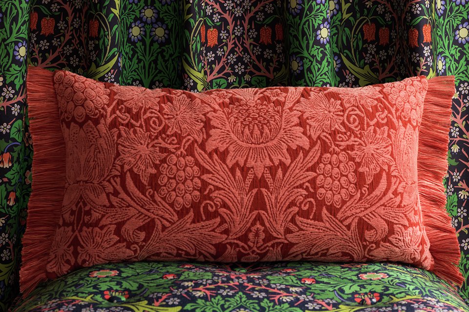 Red floral print long cushion with tuffed edge infront of green floral curtains.