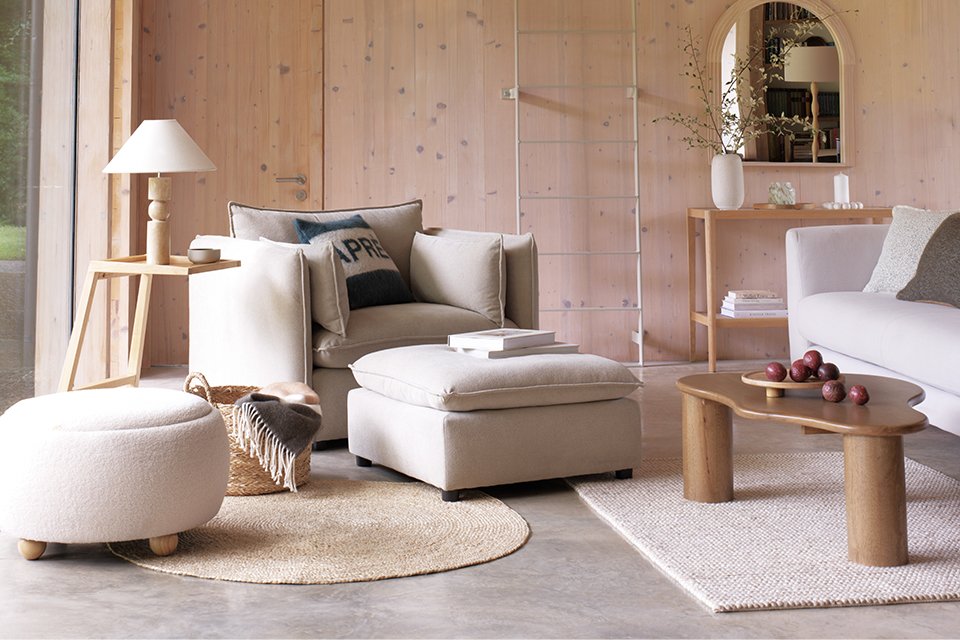Cosy modern living room with beige furniture, including a comfortable armchair, ottoman, and sofa. The room features a wooden coffee table with decorative apples, a side table with a lamp, and a wicker basket with blankets. A large mirror and vase with branches adorn the wooden wall in the background.