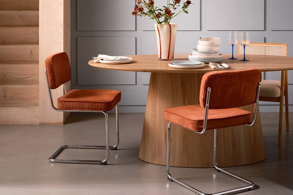 A room with orange dining chairs placed beside a wooden dining table with flowers in a vase on top of it.