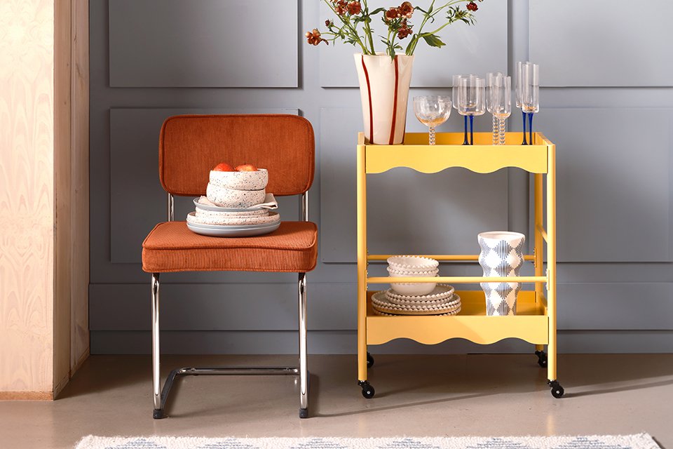 An orange chair next to a yellow trolley with plates and glasses placed on it.