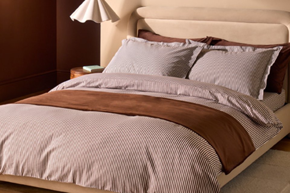 Cream bed in bedroom with stripe brown bedding and wavy white ceiling light.