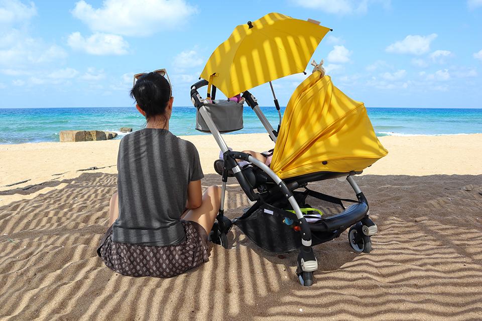 Woman sat on the beach, facing to the sea with a pushchair next to her under a pushchair parasol.