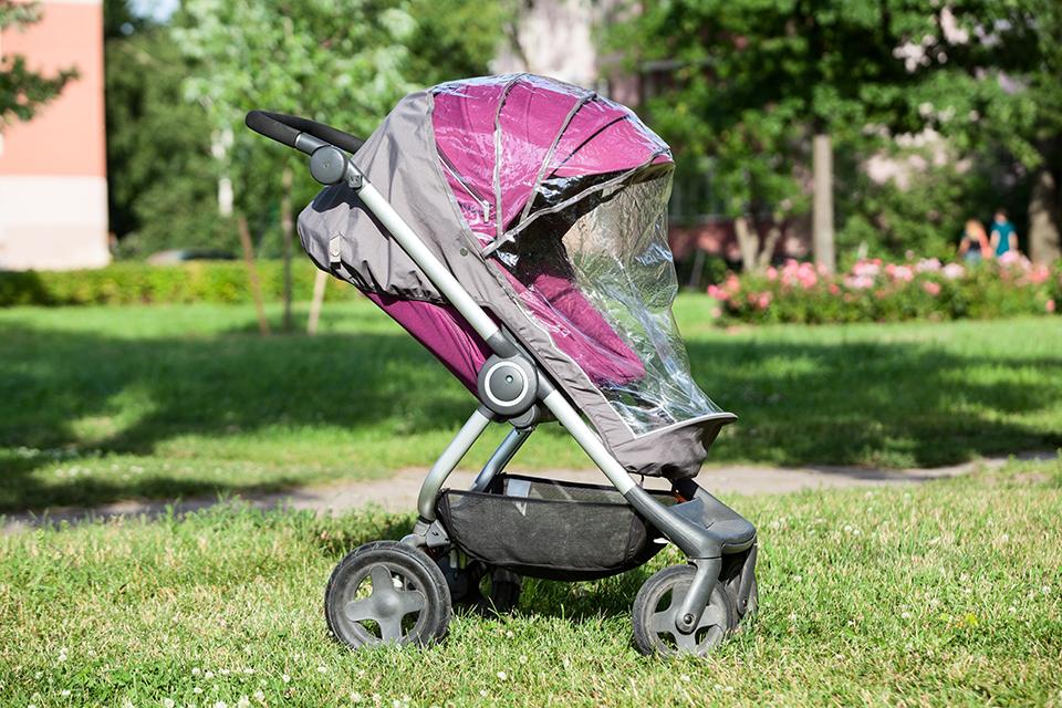 Purple pushchair in the park, with the hood up and a raincover on top.