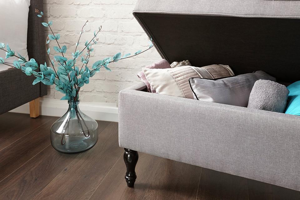 Grey ottoman with brown wooden legs, open to reveal pillows and throws inside.