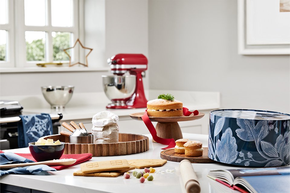 Baking essentials including cake tin, cake stand, wooden tray, rolling pin, mixing bowl etc placed on a counter in a kitchen.