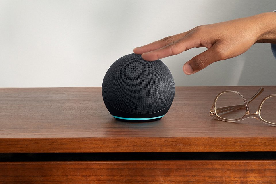 A person tapping an Echo Dot smart speaker with Alexa placed on a wooden table next to eye glasses.
