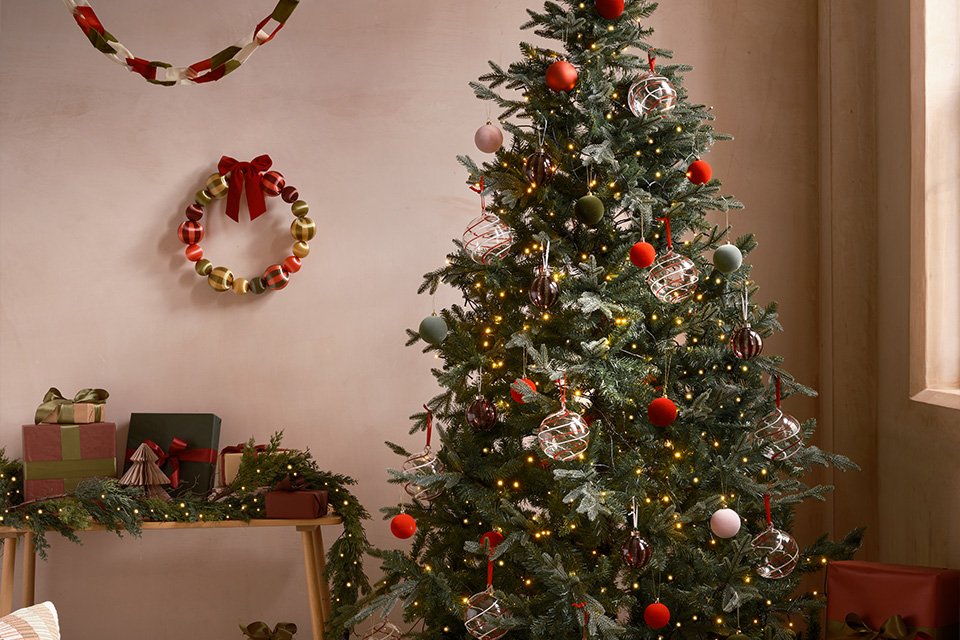 A big Christmas tree decorated with lights and baubles next to a sofa.