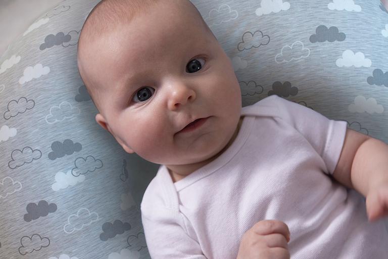 A baby girl in a pink vest laying on a Chicco grey cloud nursing pillow.