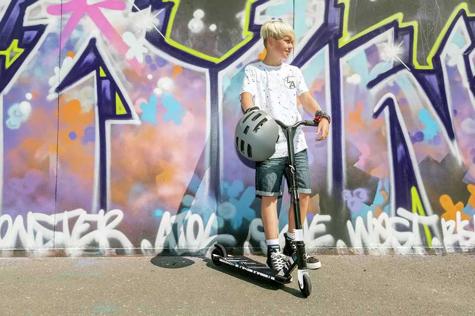 A boy on his 2 wheel scooter against a colourful wall.
