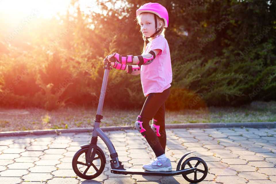 A beautiful girl in a pink protective outfit rides a scooter through a park.