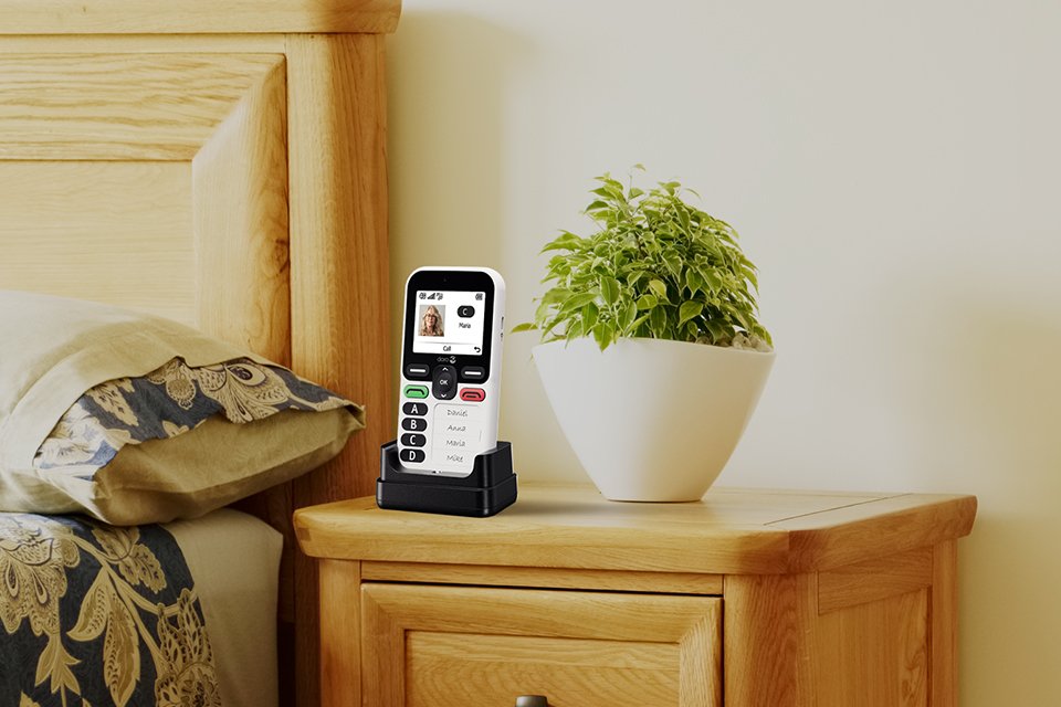 Doro Leva X10 feature phone placed in a charging cradle on a bedside table next to a potted plant.