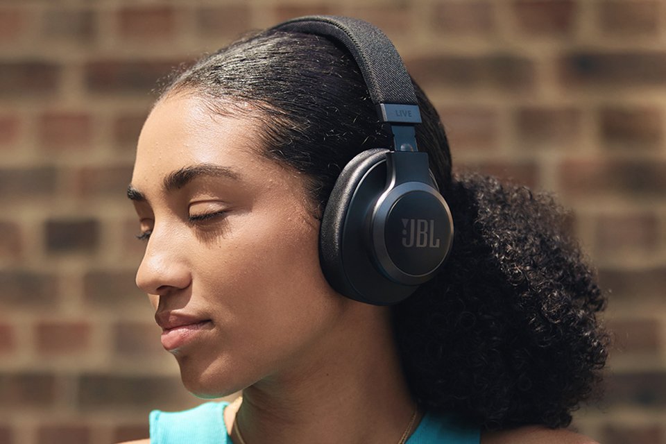 Woman wearing JBL Live 770NC headphones with brick wall background.