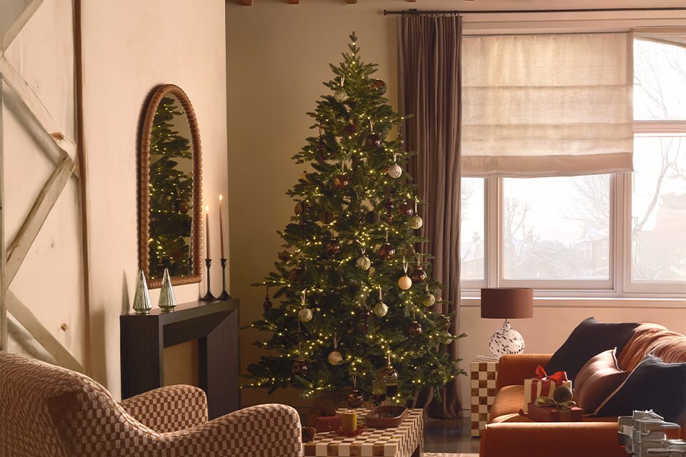 A Christmas tree decorated with lights and baubles next to a wreath and gift boxes in a living room.