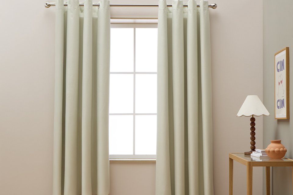 White Habitat blackout eyelet curtains drawn over a window next to a side table with a lamp on it.