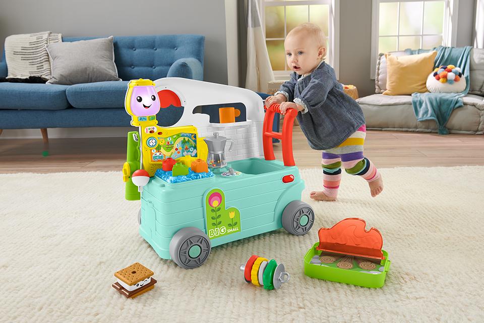 A baby is walking with the aid of a brightly-coloured Fisher-Price toy camper.