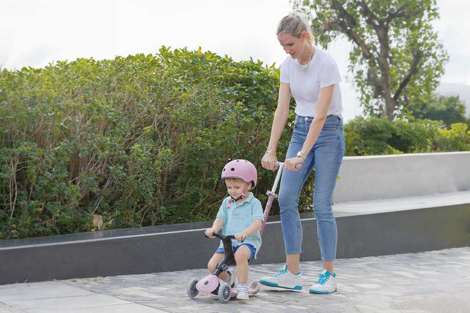 A mom helping her toddler ride a scooter.