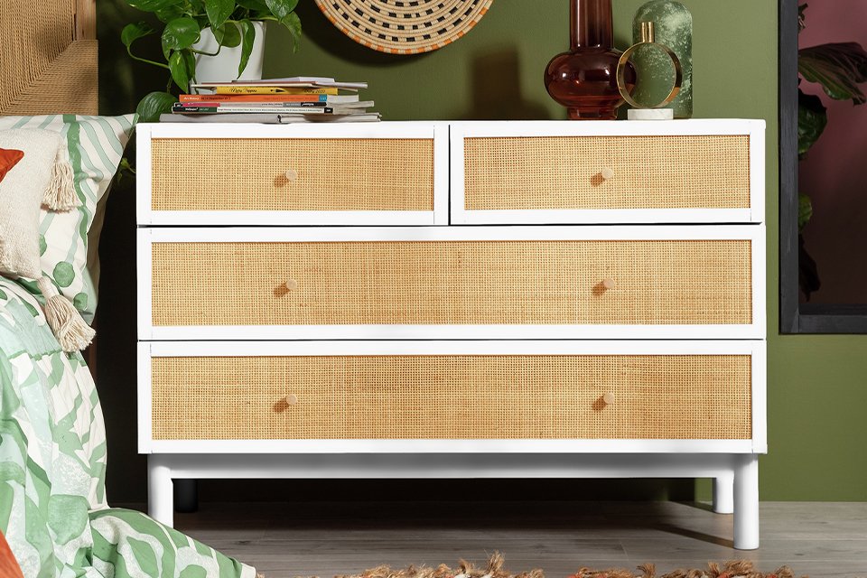 Habitat Simone Wide Rattan 4 Drawer Chest in white and natural finish placed next to a bed. 