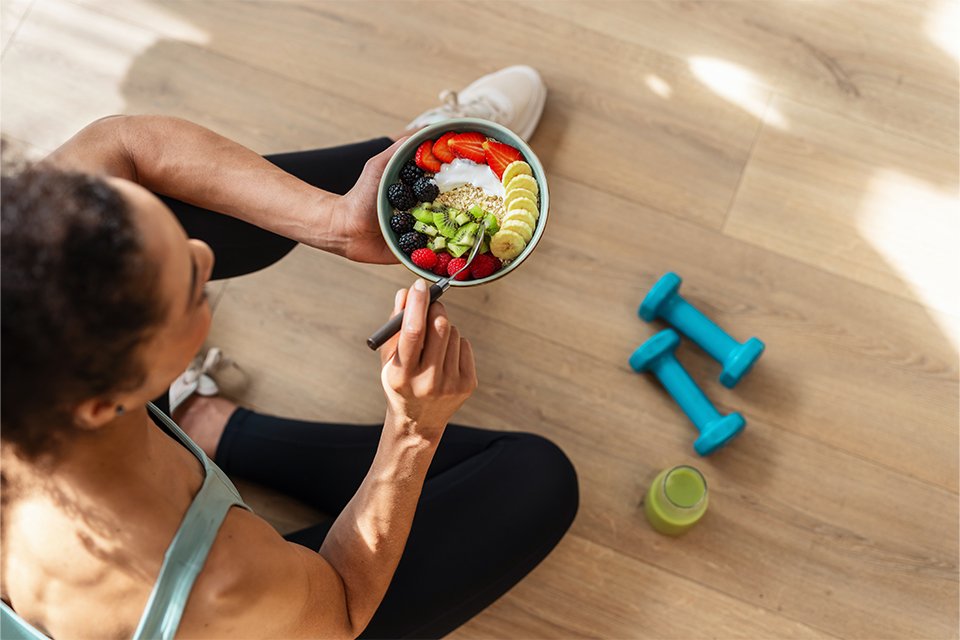 A woman having breakfast sitting on the floor with blue dumbbells and a bottle of healthy juice placed next to her.