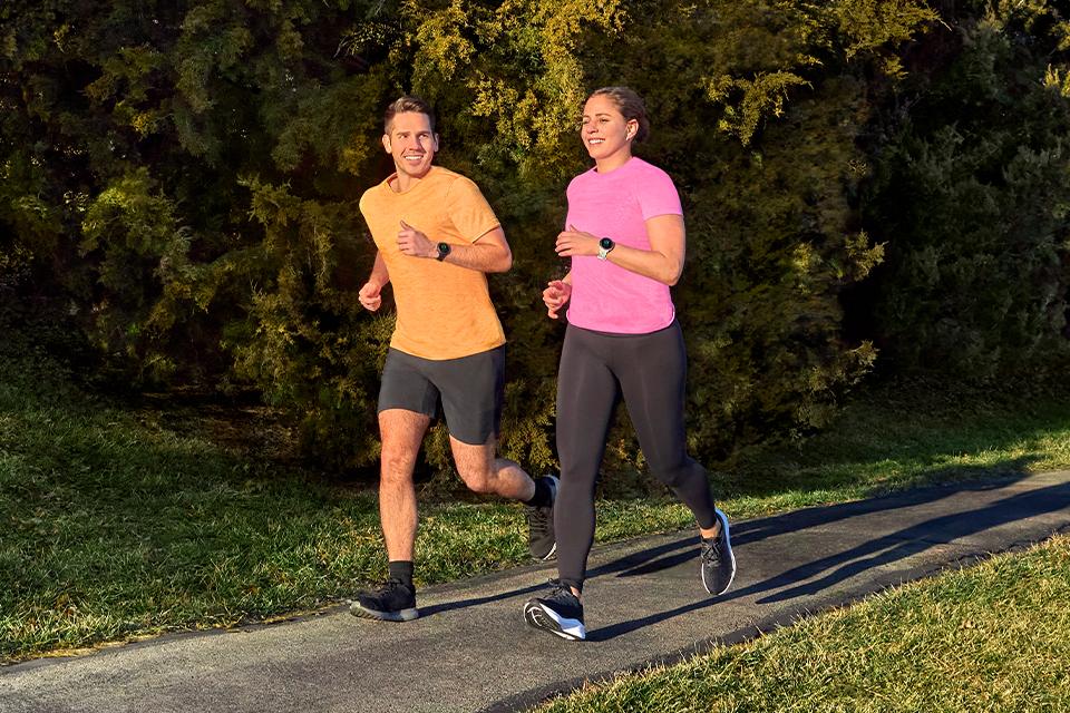 A man and a woman jog along a path in a sunny park, both wearing brightly-coloured T-shirts and Garmin Forerunner running watches.
