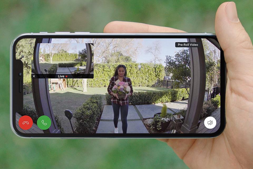 Person holding a mobile phone that is using a Ring video doorbell to monitor their front door, which is showing a woman approaching with flowers.
