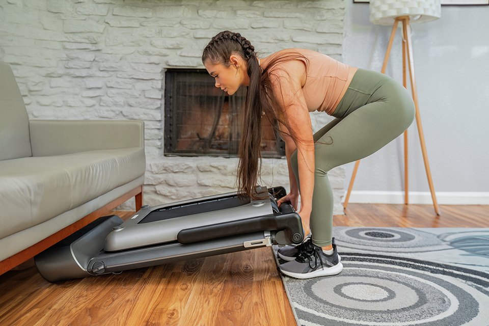 A woman storing a foldable treadmill below a sofa.