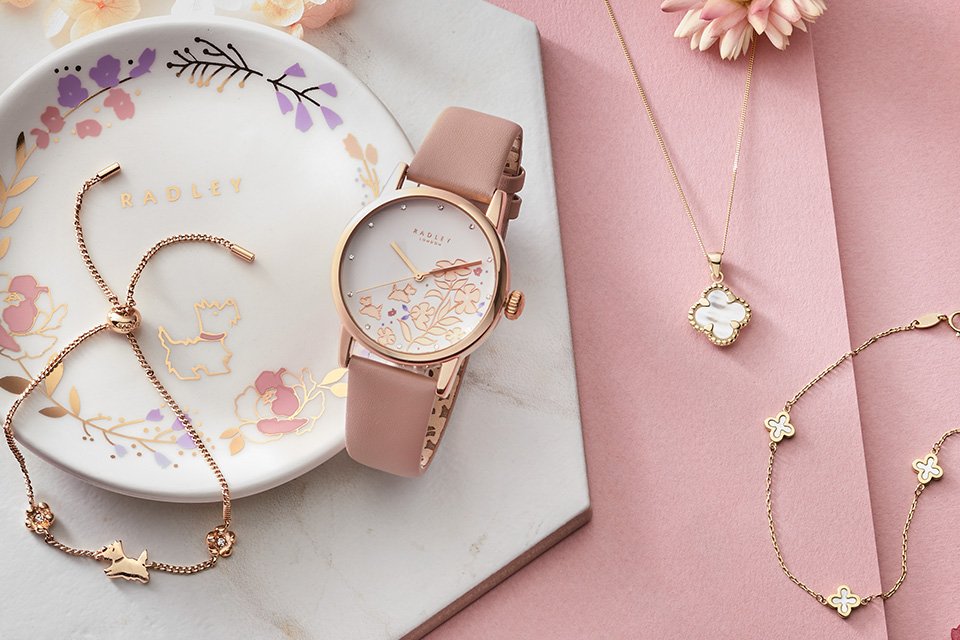 A collection of rose gold jewellery and a pink watch displayed on a pink and white background.
