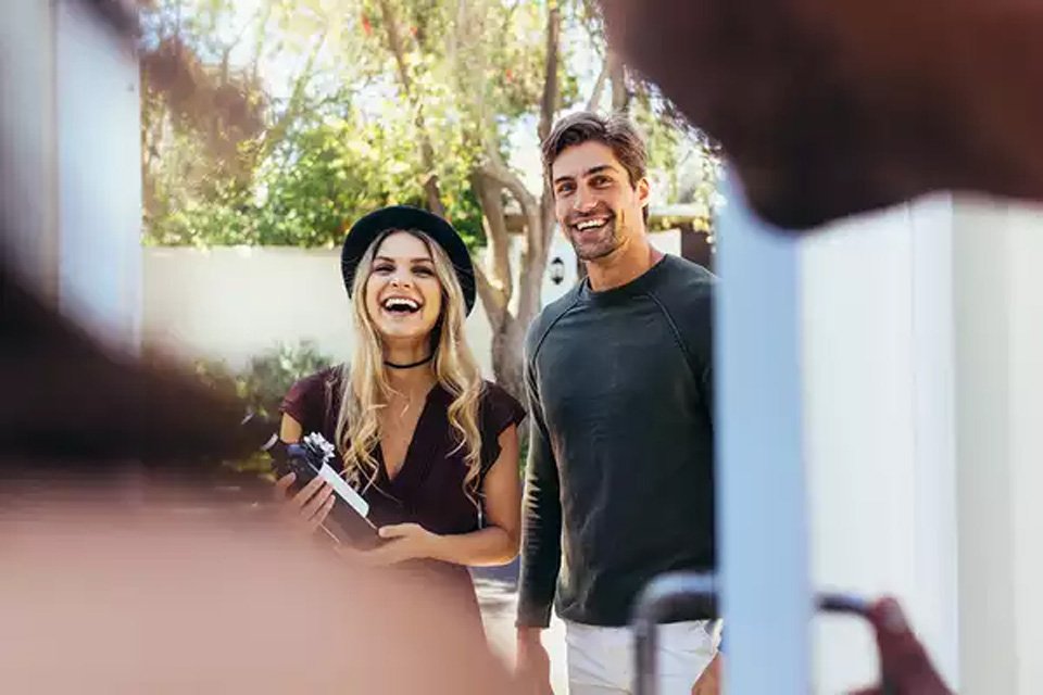 A couple entering a house with a bottle of wine.