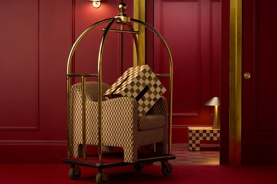 A trolley in a hotel lobby with a chequered box on top of a chequered sofa next to a table lamp on a chequered coffee table.