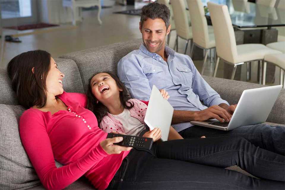 A family of 3 relaxing on a sofa watching TV and using a laptop and tablet.