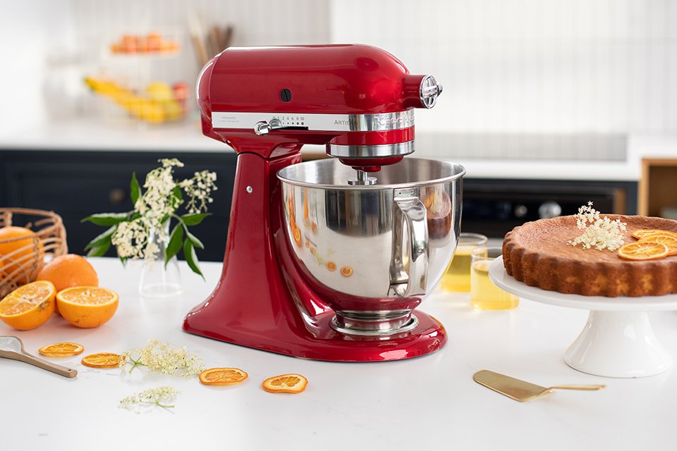 A red tilt-head KitchenAid stand mixer placed next to orange slices and an orange cake.