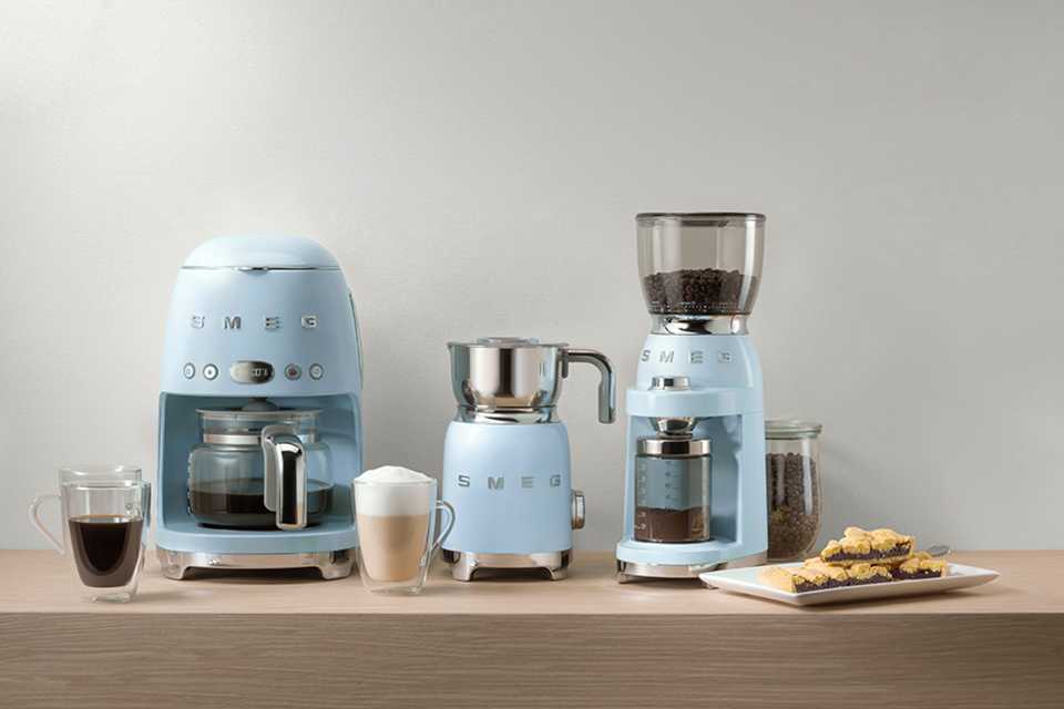 The Smeg drip filter coffee machine, coffee grinder and milk frother in pastel blue colour placed on a wooden desk.