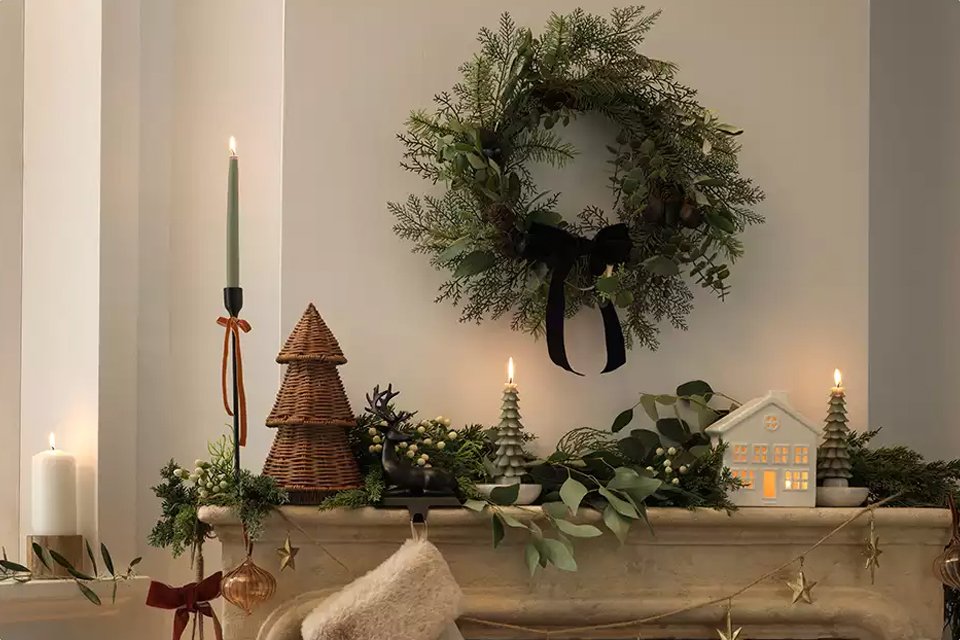Elegant Christmas mantel decor featuring a green wreath with black ribbon, candles, wicker tree ornaments, festive garland, and a glowing ceramic house.